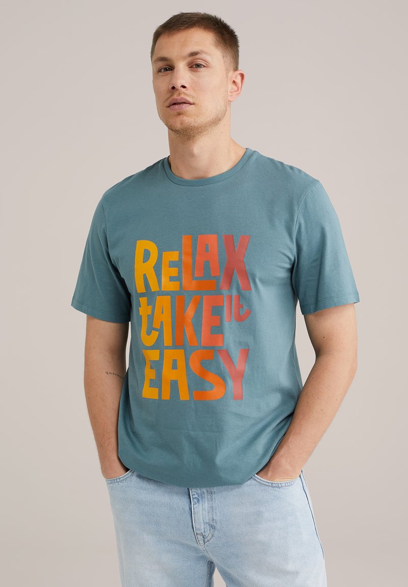 Türkiz pamut póló, amelyen merész, színes felirat található: "RELAX TAKE it EASY." Kerek nyakkal és rövid ujjakkal rendelkezik, lazább szabásban.