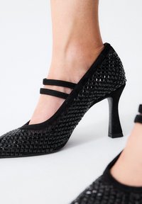 Zapatos de tacón alto negros con un patrón texturizado de tachuelas. Presentan dos correas delgadas en el empeine y una punta afilada. Fabricados con material sintético.