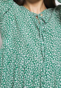 Abito verde con stampa floreale bianca, caratterizzato da un collo a chiave, maniche corte e tessuto arricciato per una silhouette fluida.