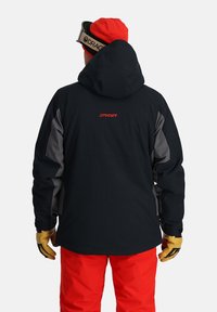 Black ski jacket with grey accents, adjustable hood, and a red logo. Paired with red snow pants and yellow gloves.
