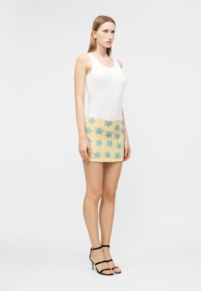 Γυναίκα που φοράει λευκό αμάνικο τοπ, μπεζ μίνι φούστα με μπλε κεντήματα λουλουδιών και μαύρα σανδάλια με ψηλό τακούνι, στέκεται μπροστά σε έναν απλό φόντο.