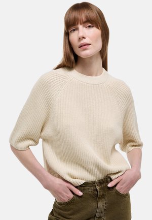 Femme portant un pull en tricot à manches courtes crème et un pantalon vert olive, les mains dans les poches, sur un fond blanc uni.