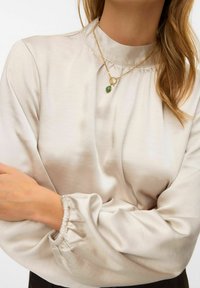 Lichtbeige satijnen blouse met een hoge kraag en geplooide manchetten. Heeft een gouden gelaagde ketting met een groene stenen accent.