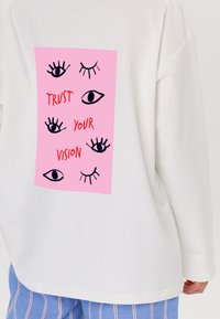 Witte lange mouw shirt met een roze graphic op de rug met verschillende oogillustraties en de woorden "VERTRAAG OP JE VISIE" in het rood.