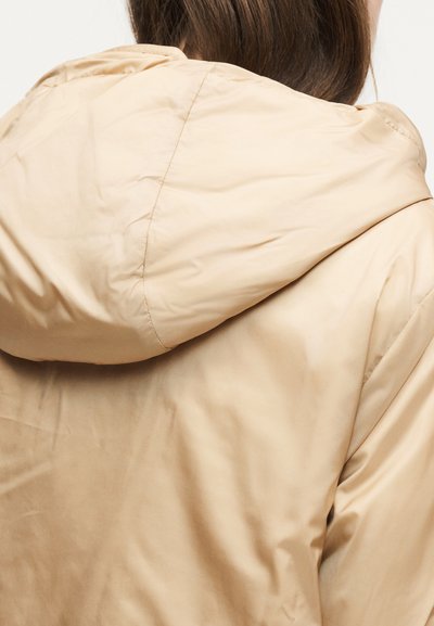 Γυναίκα με μπεζ μπουφάν με κουκούλα και εμφανείς ραφές, εμφανίζεται από πίσω με μακριά καστανά μαλλιά που είναι μερικώς ορατά.