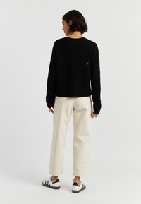 Pull à mailles noires avec des manches longues, ourlet côtelé et un petit logo en forme de cœur rouge au dos. Associé à un pantalon beige clair et des baskets à rayures noires.