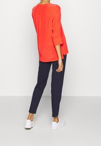 Donna che indossa un ampio top rosso brillante a maniche lunghe, pantaloni tapered di un blu navy scuro e sneakers bianche, in piedi su uno sfondo bianco semplice.