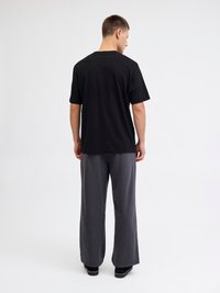 Mann steht mit dem Rücken zur Kamera, trägt ein schwarzes Kurzarmhemd und lockere graue Hosen vor einem schlichten weißen Hintergrund.