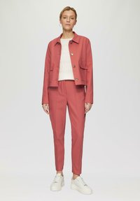 Koraalkleurige cropped jas met voorzakken en zilveren knopen, gecombineerd met bijpassende broeken. Gestructureerd wit sweater en stevige witte sneakers.