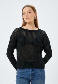 Svart stickad tröja med ribbad struktur, har en lös passform, vid nacköppning och något genomskinlig design. Tillsammans med blå jeans.