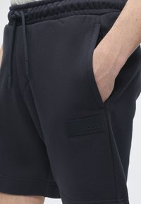 Κοντινό πλάνο ενός ατόμου που φοράει μαύρο σορτς με ελαστική μέση, κορδόνι, πλαϊνή τσέπη και μαύρο πατς με το λογότυπο BOSS.