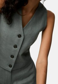 Ärmelose graue Weste mit vier schwarzen Knöpfen, getragen von einer Person mit mittlerem Hautton und lockigem, dunklem Haar.