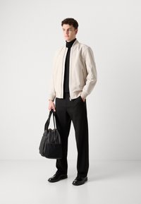 Helebeežist bomber-jakk, millel on lukuga kinnitamine, must turtleneck, mustad püksid ja must kott, seistes ühtlase tausta ees.