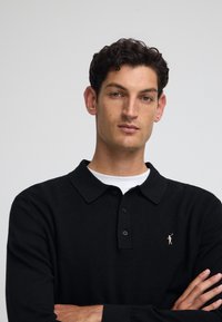 Svart pikétröja i strukturerad tyg, med klassisk krage, treknapplåttning och en liten rosa logotyp på bröstet.