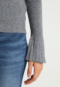 Närbild av en persons hand som bär en grå räfflad tröja med utsvängda ärmar och blå denimjeans.