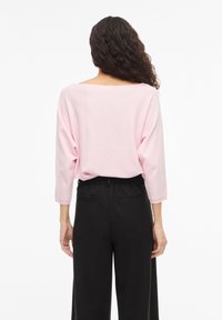 Sieviete ar gariem cirtainiem matiem, valkājot gaiši rozā vaļīgu džemperi un melnas augsta vidukļa bikses, stāvot ar muguru pret baltu fonu.