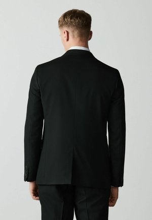 Mann mit kurzen Haaren, der eine schwarze Anzugjacke und Hose trägt, steht mit dem Rücken zu einem schlichten hellen Hintergrund.