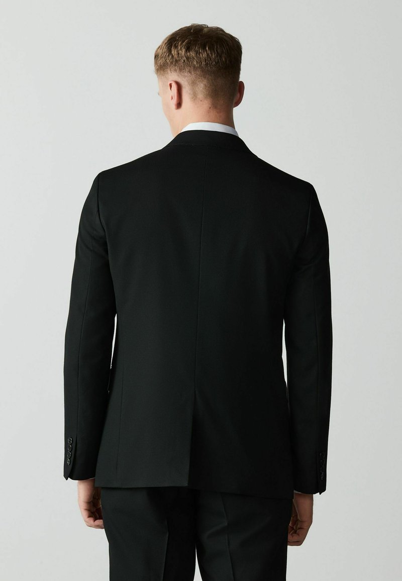 Homme aux cheveux courts portant une veste et un pantalon de costume noirs, debout de dos sur un fond clair uni.