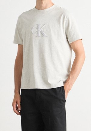 Man draagt een lichtgrijs Calvin Klein Jeans T-shirt en zwarte broek, met één hand in de zak, tegen een effen witte achtergrond.