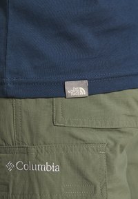 Camiseta de algodón marina con una etiqueta gris visible de "The North Face", complementada por pantalones cortos cargo oliva con un logo blanco de "Columbia" y bolsillos.