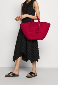 Kobieta w czarnej, teksturowanej czarnej sukience midi bez rękawów oraz czarnych sandałach na platformie, trzymająca dużą czerwoną plecioną torbę z frędzlami.
