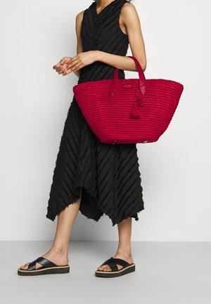 Kvinde iført en ærmeløs sort tekstureret midikjole og sorte platform sandaler, mens hun holder en stor rød vævet tote taske med frynser.