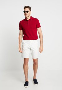 Röd polo skjorta, vita shorts, marinblå slip-on skor och svarta solglasögon. Slät tyg med krage och liten logga på bröstet.