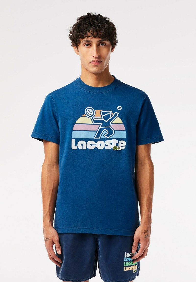 Camiseta de algodón azul con un diseño gráfico que muestra a un tenista y franjas de arcoíris sobre el logo de "Lacoste". Cuello redondo, mangas cortas.
