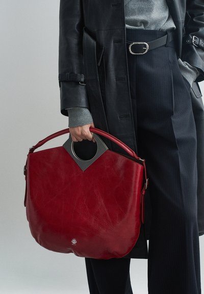 Sac à main en cuir rouge avec une poignée arrondie et un accent triangulaire gris. Le sac présente une surface texturée et est tenu par une personne.