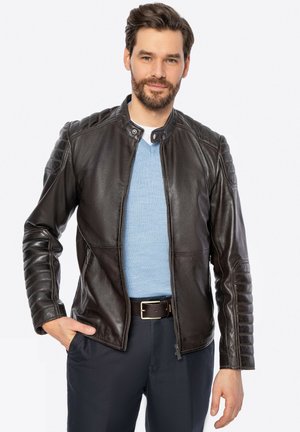 Zwarte leren jas met een staande kraag, gequiltte schouderaccenten en een ritsluiting. Gedragen over een lichtblauw shirt.