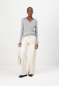 Hellgrauer langärmliger V-Ausschnitt-Pullover, cremefarbene weit geschnittene Hose und schwarze flache Schuhe. Das Model hält eine beige Tasche mit strukturiertem Design.