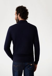 Maglione blu navy con colletto e polsini a costine, realizzato in tessuto liscio. La vista posteriore mostra un design aderente abbinato a jeans in denim blu.