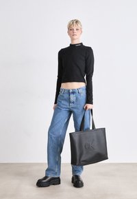 Haut noir à manches longues cropped, jeans bleu clair taille haute, sac tote en cuir noir avec logo "CK" en relief, bottes noires chunky.