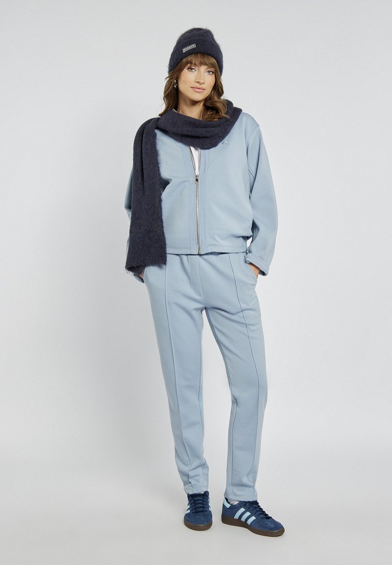 Jasnoniebieska bluza zapinana na zamek oraz dopasowane spodnie z miękkiego materiału, w połączeniu z ciemnoniebieskim szalikiem i czapką. Granatowe trampki Adidas z jasnymi paskami.