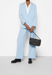 Traje ajustado de color azul claro con una blazer de un solo botón y pantalones de pierna ancha. Bolso negro bandolera con un patrón texturizado y una correa corta. Botas negras de suela gruesa.