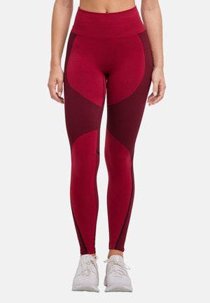 Kobieta nosząca legginsy z wysokim stanem w czerwonym i ciemnoczerwonym wzorze, w połączeniu z białymi butami sportowymi, stojąca na białym tle.