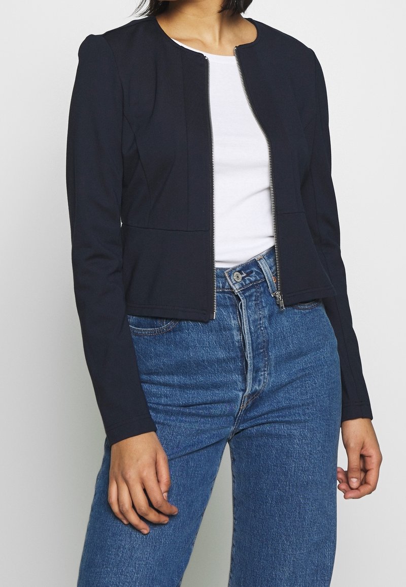 Femme portant une veste noire zippée par-dessus une chemise blanche associée à un jean bleu taille haute sur un fond clair uni.