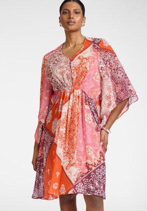 Kobieta w wielokolorowej pomarańczowo-różowo-fioletowej sukience patchworkowej w kwiaty z rękawami do łokcia i srebrnymi kolczykami kołami.