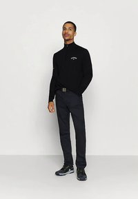 Άνδρας που στέκεται με μαύρο πουλόβερ quarter-zip, σκούρα παντελόνια και μαύρα αθλητικά παπούτσια, σε απλό ανοιχτό φόντο.