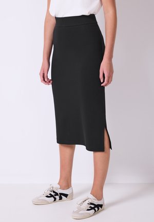 Zwarte midi-jurk met een hoge taille en zijsplits, gemaakt van rekbare stof. Gepaard met witte sneakers met zwarte accenten.