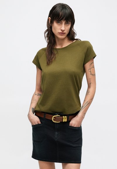 T-shirt à manches courtes vert olive, coupe décontractée, associée à une jupe en denim foncé. Ceinture marron avec boucle dorée, avec poches avant visibles.