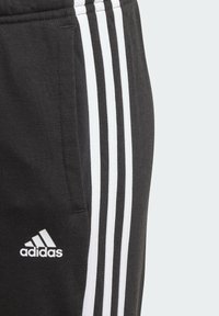 Pantalones de chándal negros con tres franjas blancas a lo largo de los lados y un logo de Adidas bordado en blanco cerca del bolsillo. Material de algodón suave.