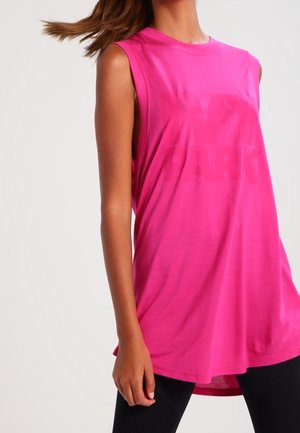 Kvinde iført ærmeløs, løs top i klart pink og sorte bukser, stående med håret let blæsende til siden mod en ensfarvet baggrund.