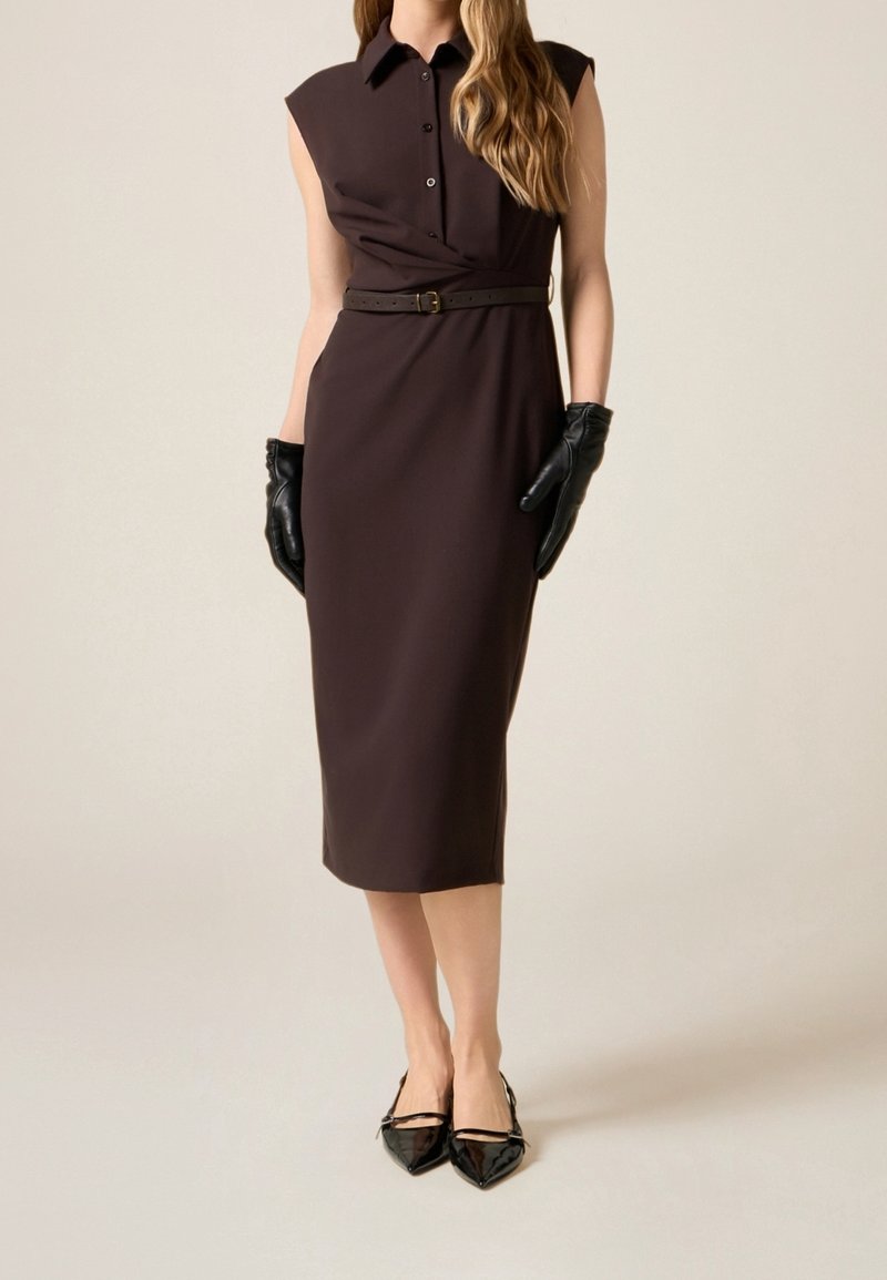 Femme portant une robe midi sans manches marron foncé avec ceinture, gants en cuir noirs et ballerines noires pointues, debout devant un fond uni.