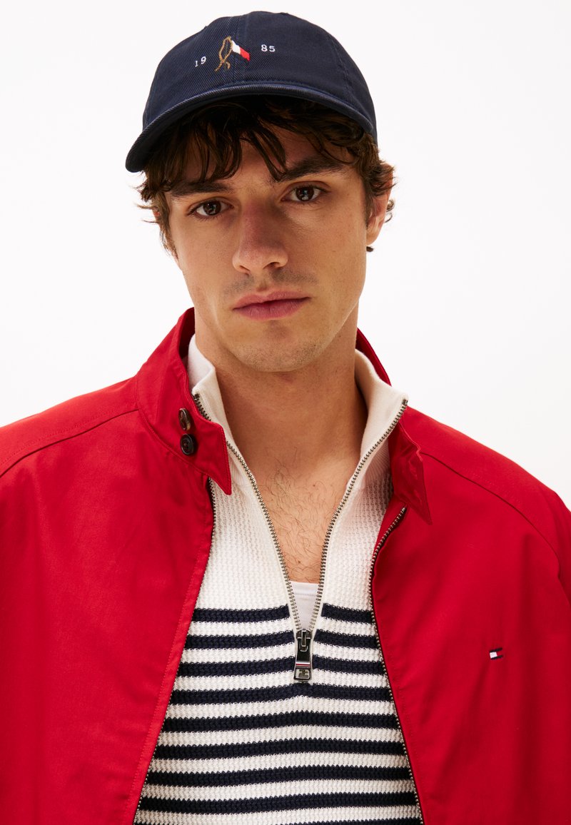 Veste zippée rouge par-dessus un t-shirt en tricot à rayures blanches et navy. Casquette navy avec logo. Cheveux courts bruns et fond neutre.
