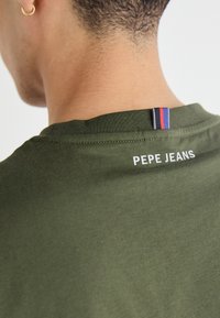 Olīvu zaļš kokvilnas T-krekls ar apaļu kaklu, uz kura ir balts "PEPE JEANS" logs un daudzkrāsaina birka aizmugurē pie kakla.