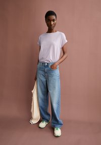 Maglietta corta a maniche corte color lavanda chiaro, jeans a vita alta blu chiaro, sneakers bianche e verdi, tiene in mano un cardigan crema. Sfondo piatto.