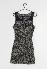 Robe noire à fleurs avec des marguerites blanches, sans manches, panneau arrière en dentelle, taille cintrée et jupe évasée. Suspendue à un crochet noir.