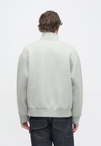 Felpa grigio chiaro con collo alto, spalle scese e vestibilità comoda. Presenta polsini e orlo a coste. Indossata sopra jeans scuri.