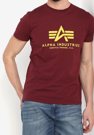 Mand iført en mørkerød t-shirt med gul Alpha Industries-logo og tekst, kombineret med blå denimjeans, stående foran en ensfarvet baggrund.